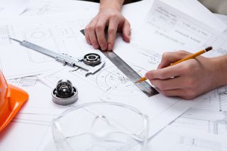 Close-up of hands using a ruler and pencil on mechanical blueprints with ball bearings, a caliper, and safety goggles.