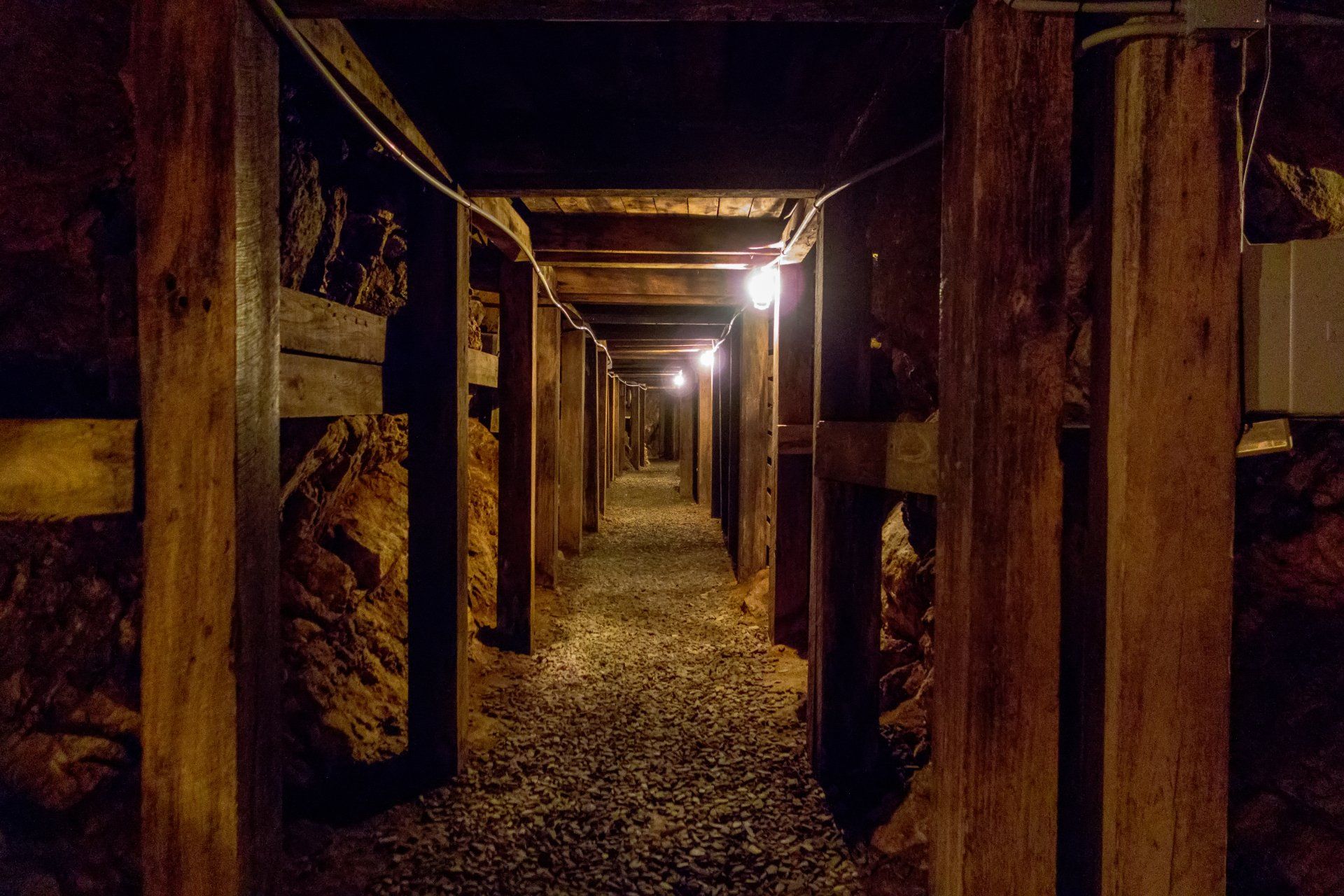 A dimly lit, narrow mine tunnel reinforced with wooden beams, featuring a gravel floor leading into the darkness.