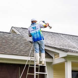 A man is standing on a ladder cleaning the roof of a house.