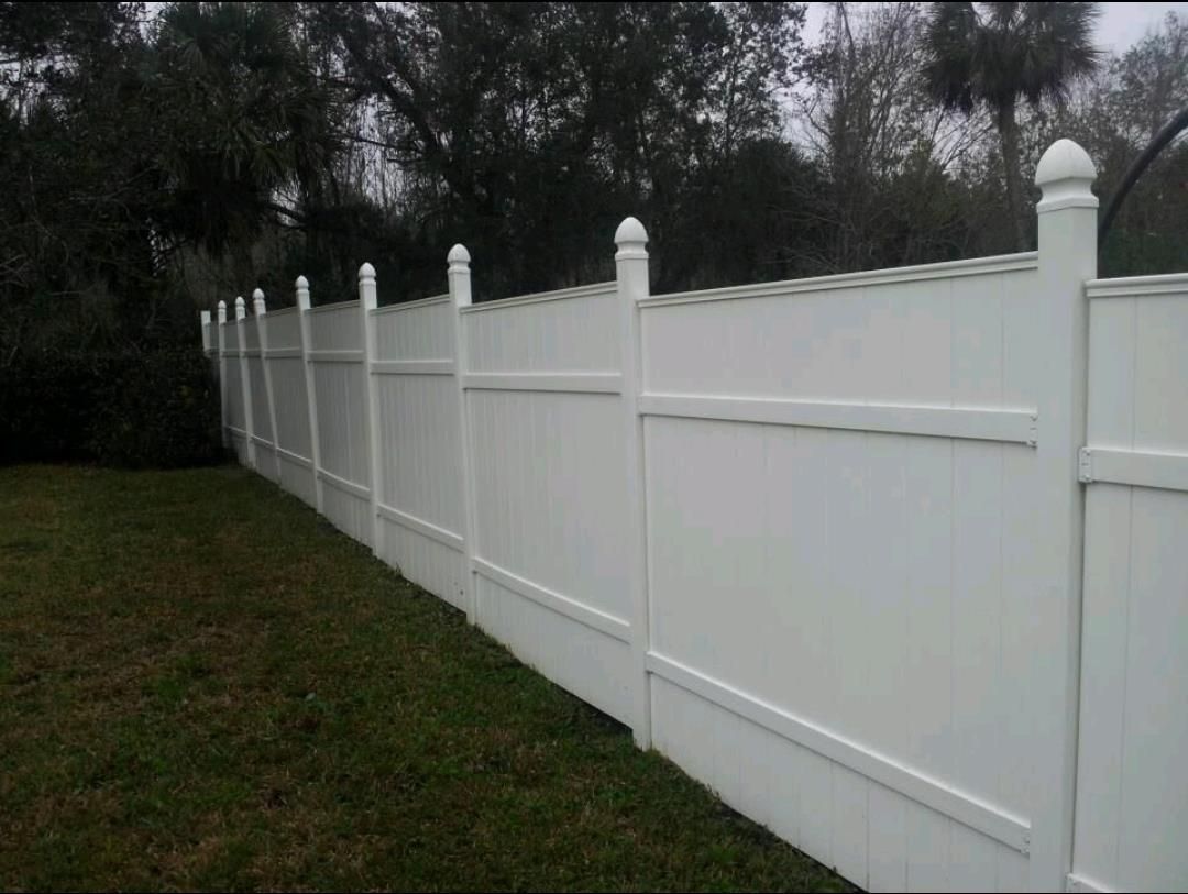 A white fence surrounds a lush green field with trees in the background.