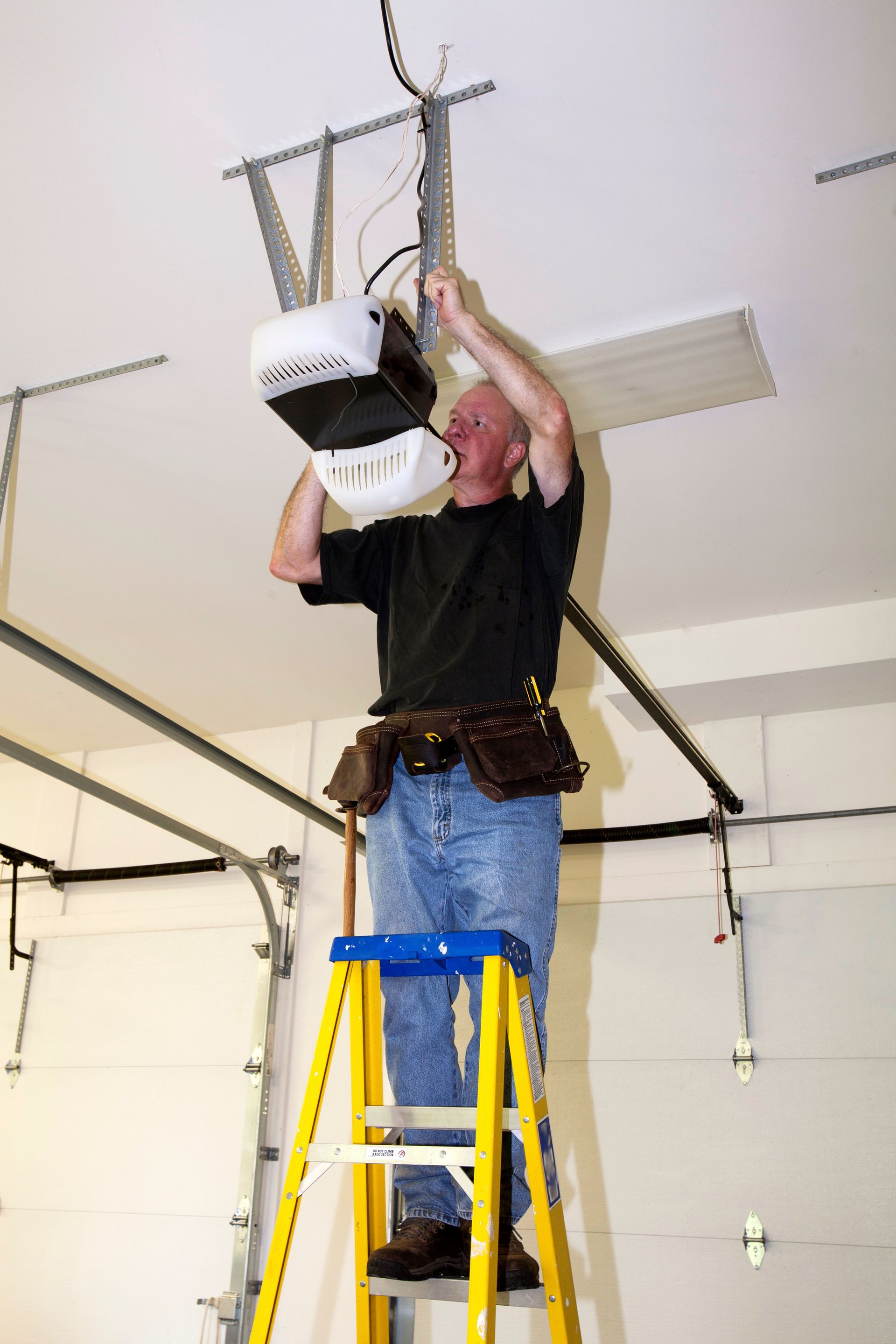 View of a modern garage door in an elegant suburban home, after a garage door installation service. A technician on a ladder is installing a garage door opener in a garage.