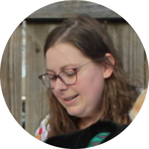 Woman with glasses looking down, smiling slightly, in front of a wooden fence.