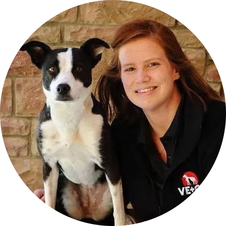 Woman in black jacket with dog, both looking forward. Brick background.