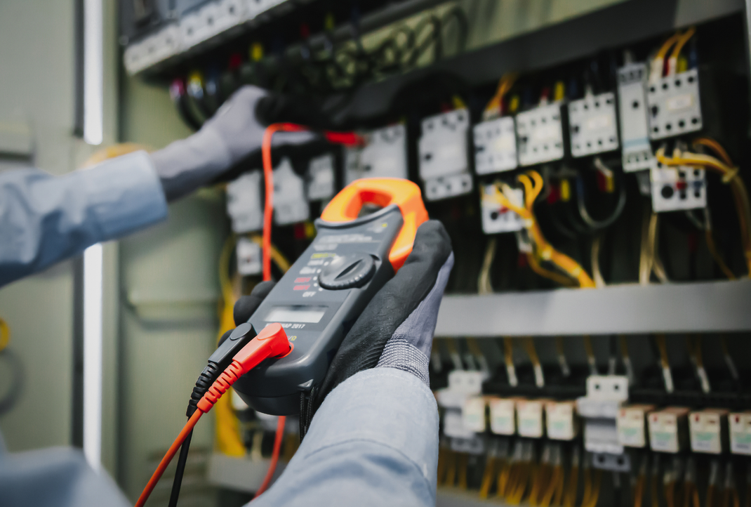 An electrician wearing gloves uses a digital clamp meter to test electrical components in a control panel.