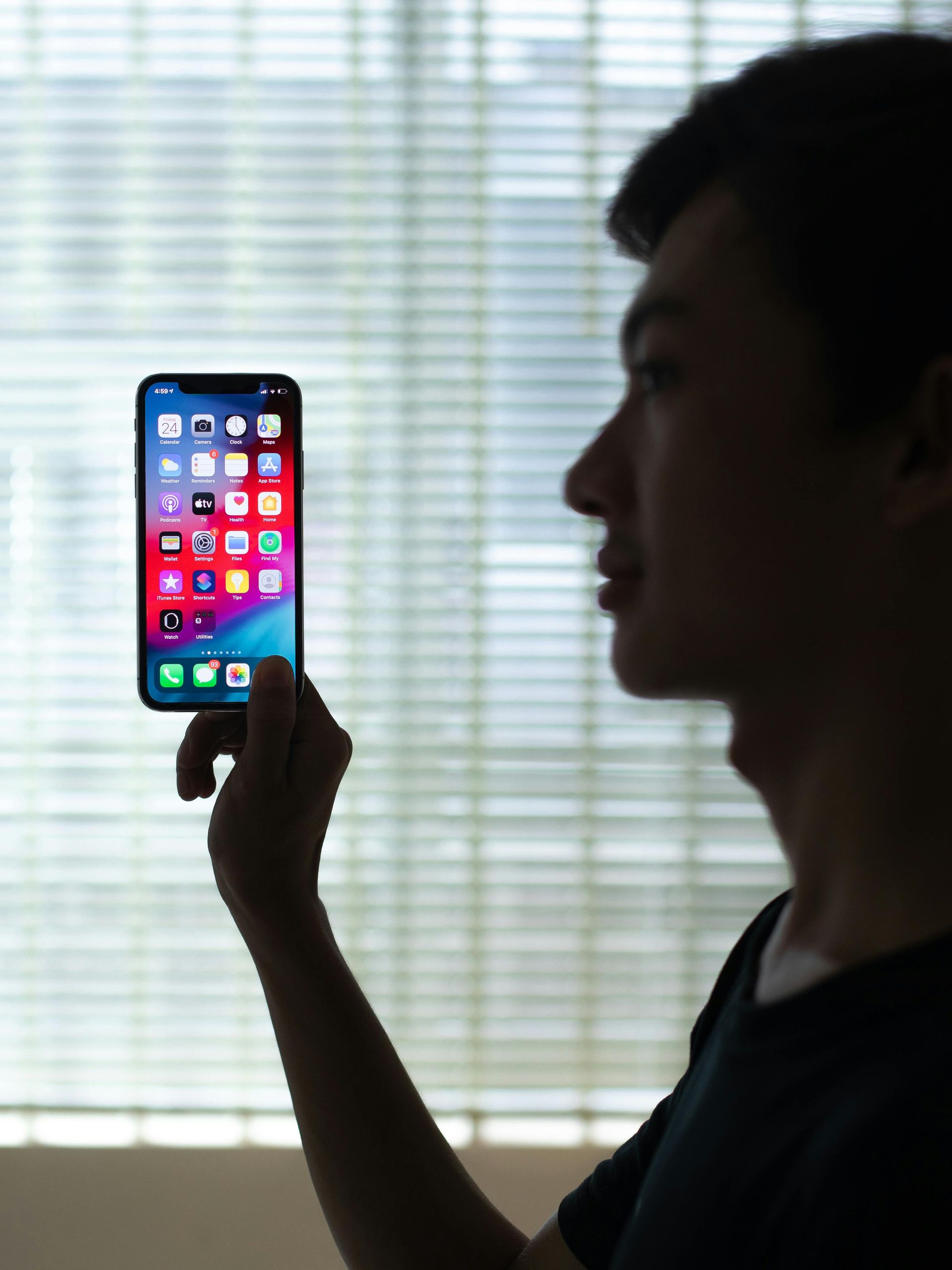 Man silhouetted holding a smartphone up to his face, in front of a window with blinds.