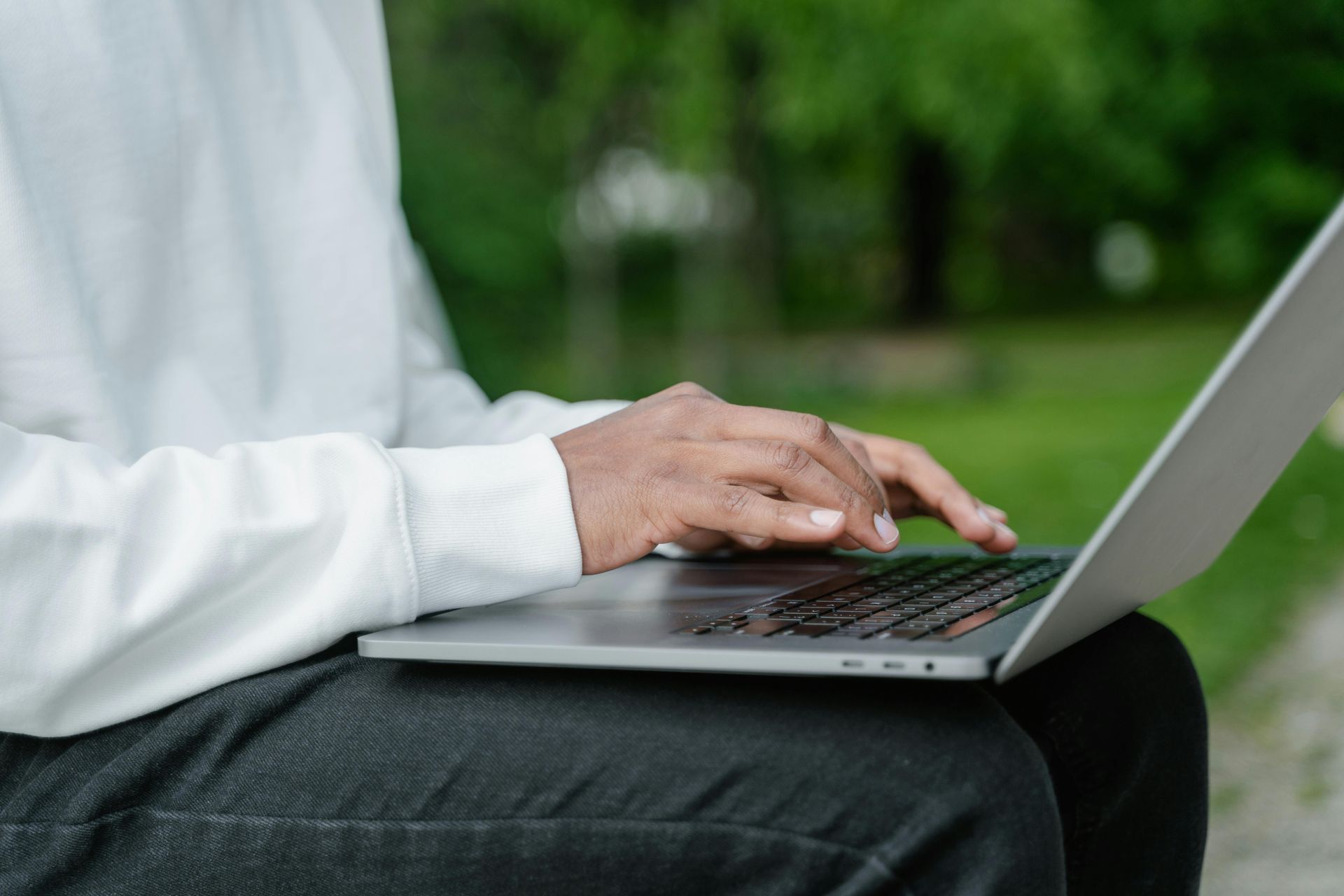 Person's hands typing on a laptop, resting on their lap. They wear a white sweatshirt, outdoors in a park.