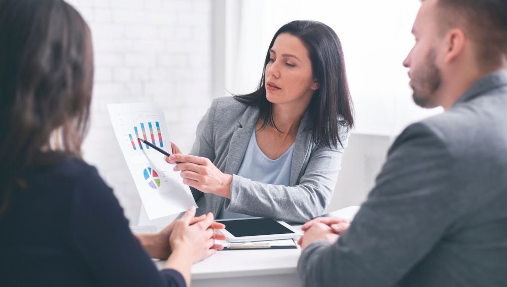 A Woman Is Giving a Presentation to A Group of People While Sitting at A Table — TJL Business Advisors & Accountants in Forster, NSW