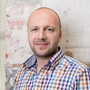 A person with a shaved head and light beard wearing a multi-colored checkered collared shirt against a brick wall.