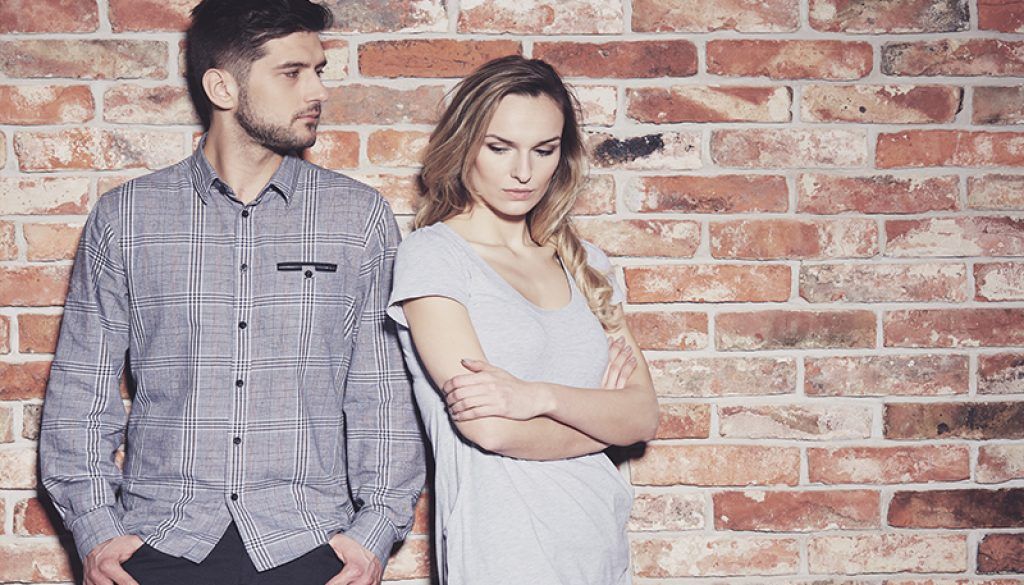 A man and woman stand against a brick wall, looking away from each other with somber expressions.