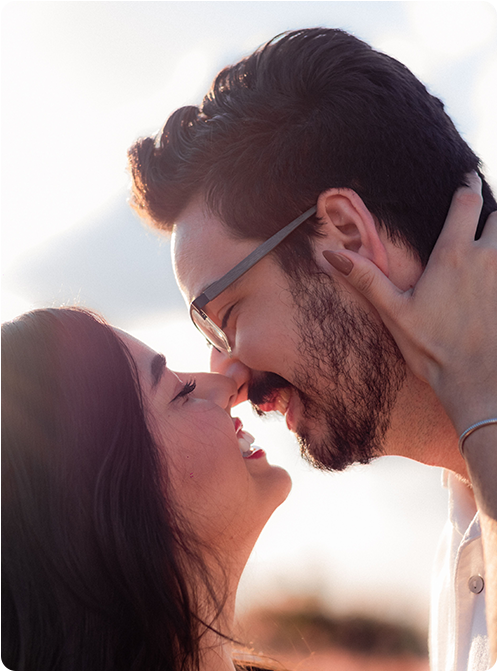 A smiling couple hugging outdoors on a sunny day, with the person in front wearing a red top and the one behind in blue.