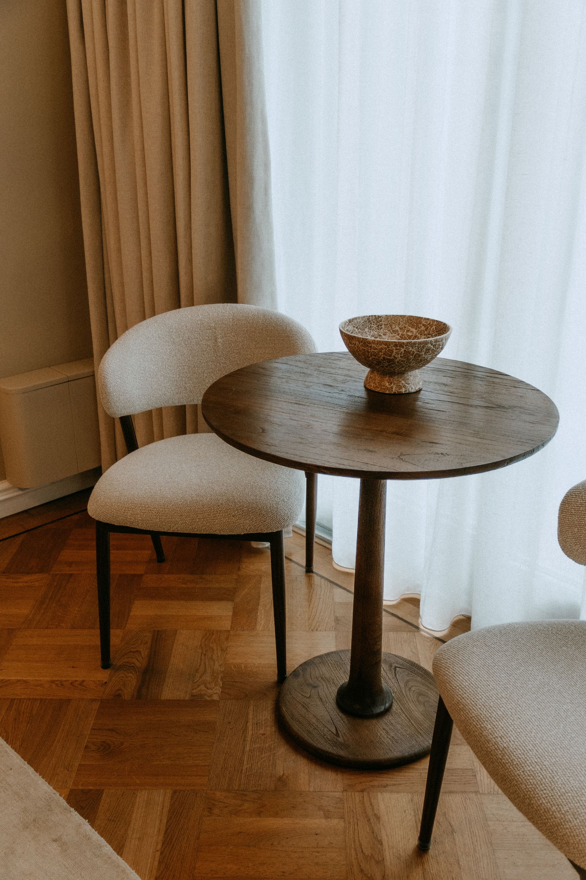Ronde houten tafel met een gevlochten schaal, geflankeerd door twee gestoffeerde stoelen in een lichte kamer met gordijnen.