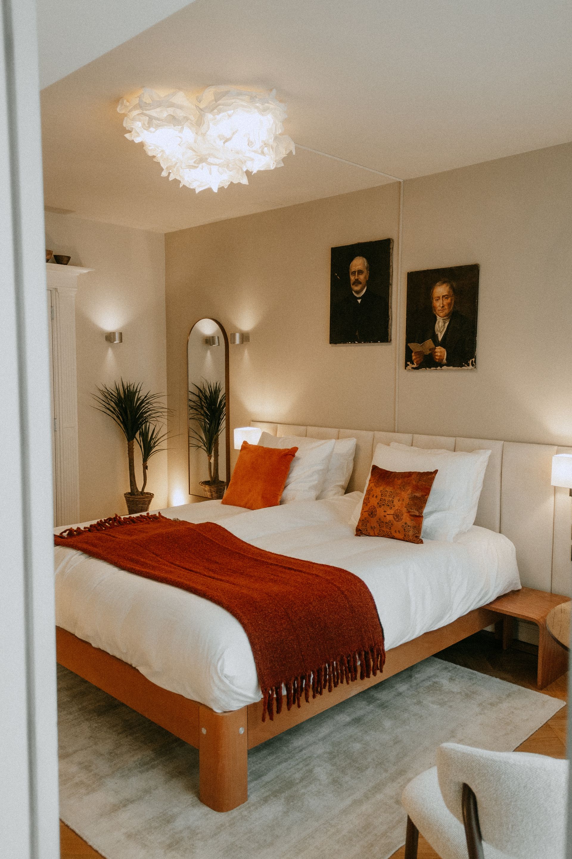 Gezellige slaapkamer met wit beddengoed, oranje accenten, ingelijste portretten en een kroonluchterachtige plafondlamp.
