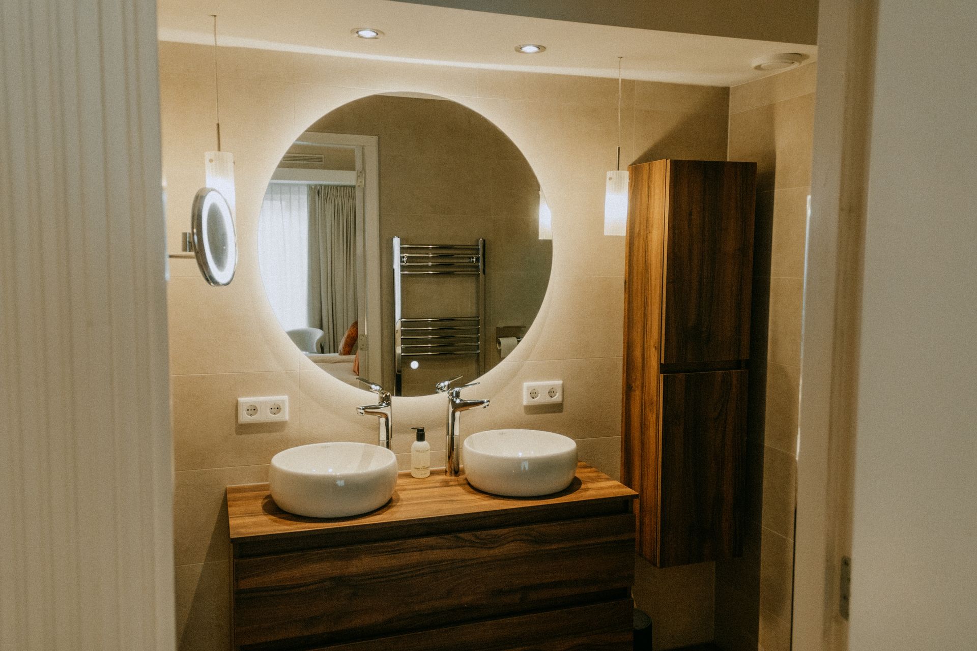 Moderne badkamerkast met ronde spiegel, dubbele wastafel en houten kastverlichting.