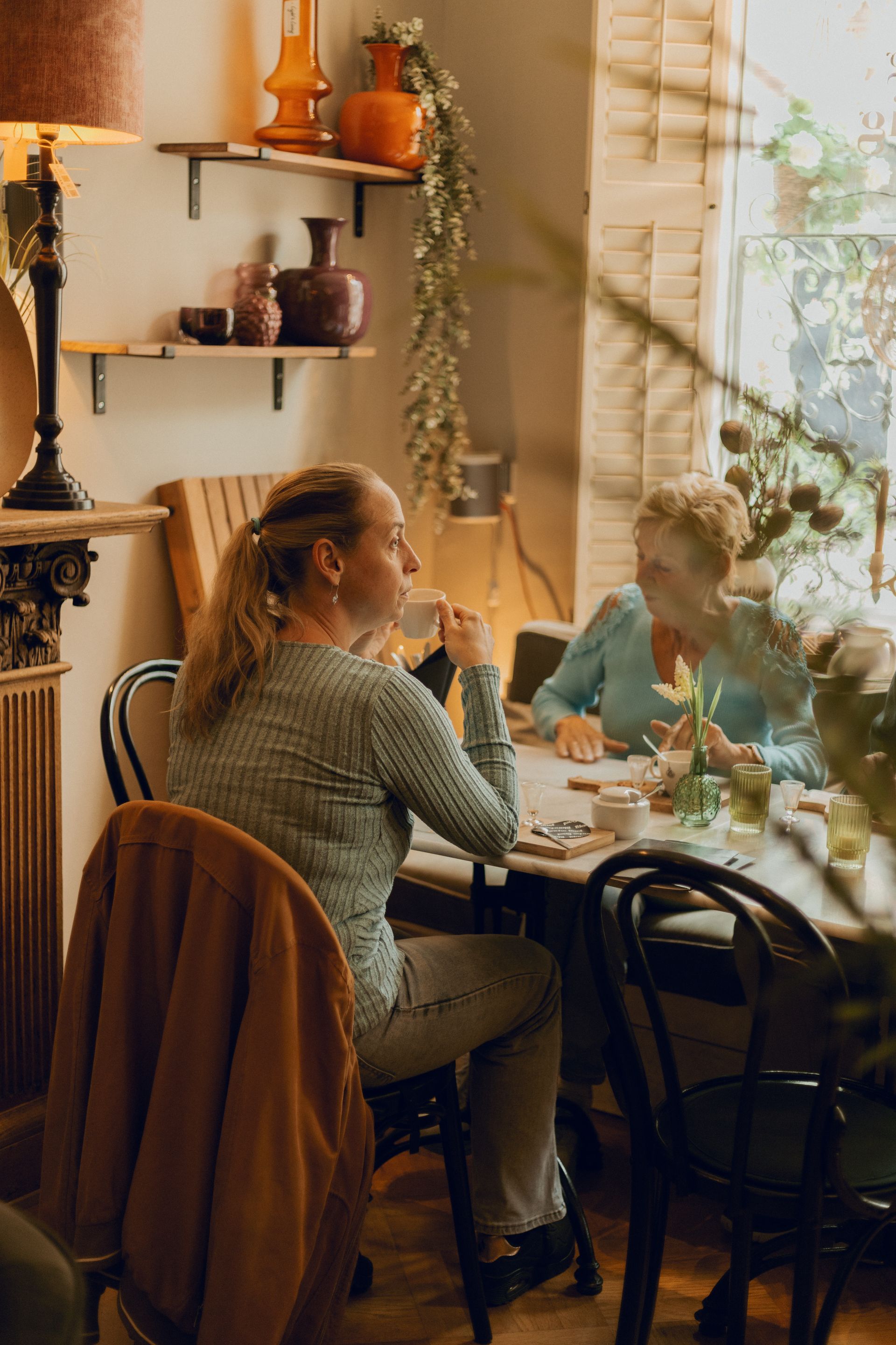 Een persoon zit aan een tafeltje bij het raam in een café, kijkend naar een spiegel met warm zonlicht en planten in de buurt.