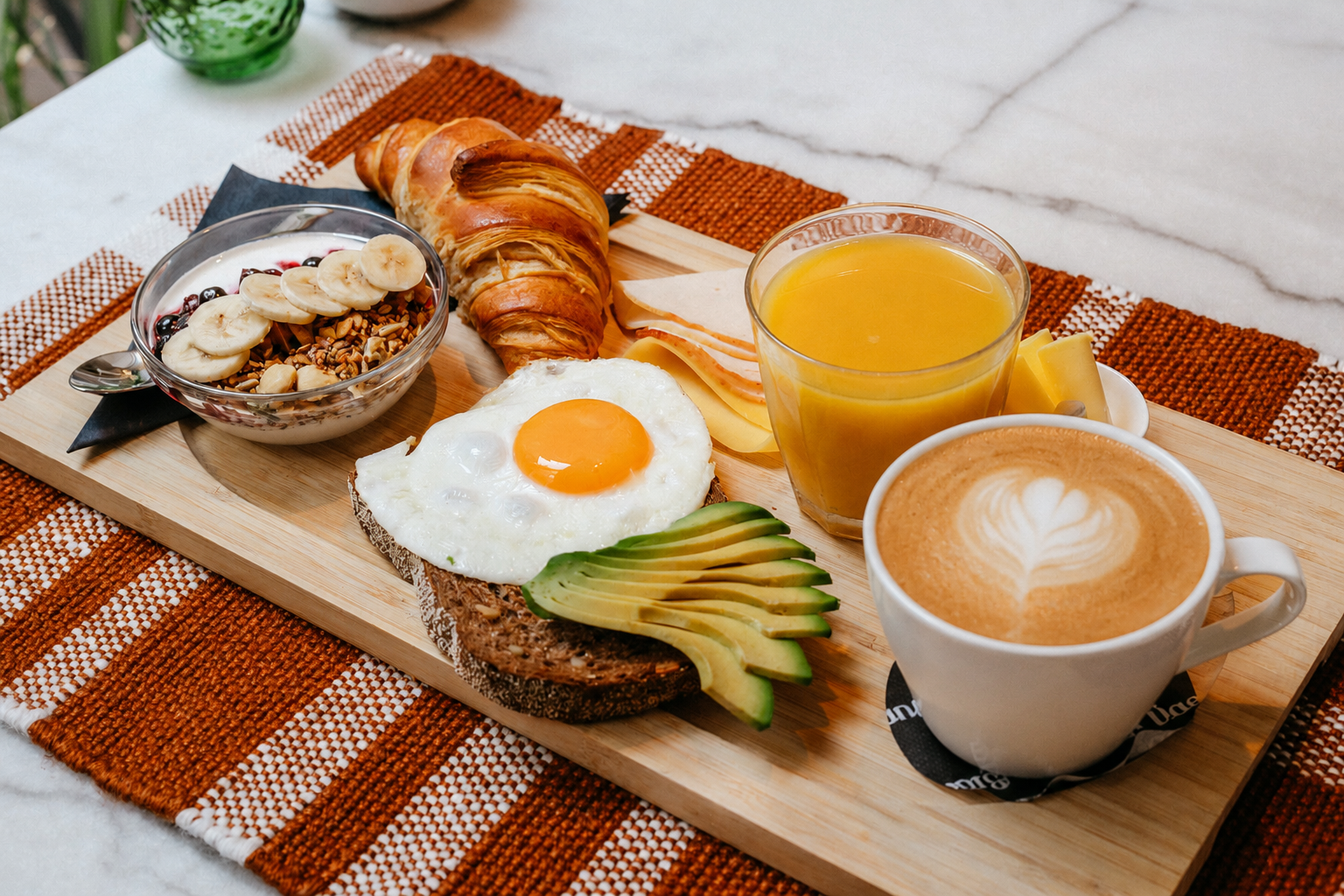 Ontbijtplateau met croissants, eieren, avocado, een fruitschaal, sinaasappelsap en latte op een geweven placemat.