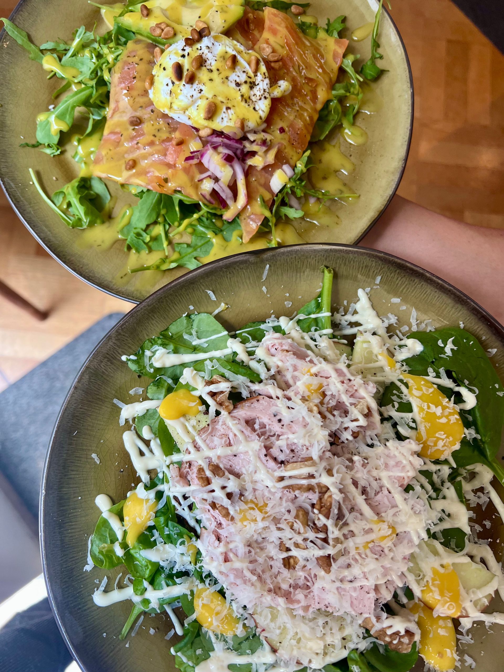 Twee salades in kommen met groene bladgroenten, mango, geraspte kaas en gegrilde kip op een houten tafel.