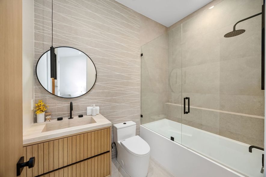 Modern beige bathroom with round mirror, wood vanity, toilet, and glass shower tub combo