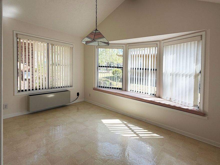 Empty beige living room with large corner windows and vertical blinds, sunlight on tiled floor.