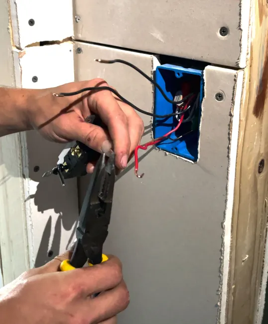 Hands wiring an electrical outlet box in a wall, using red and black wires and a wire stripper tool.