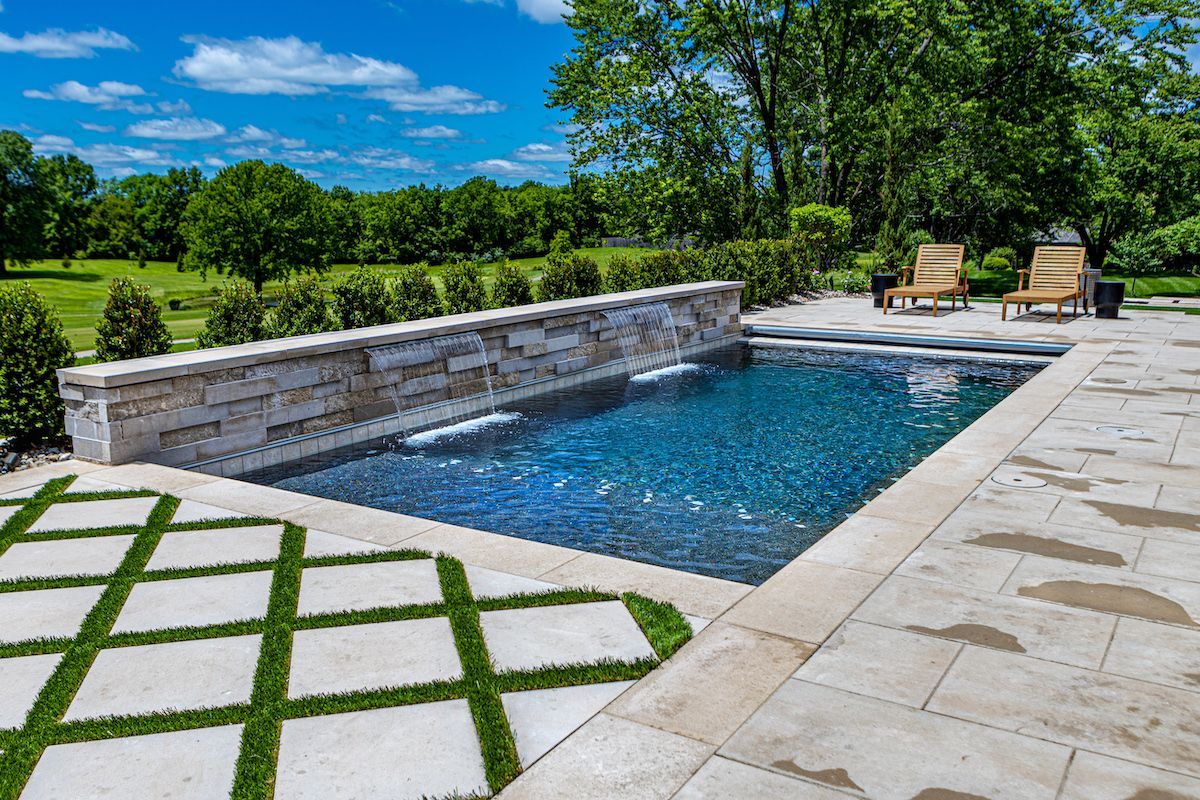 Modern backyard pool with stone deck, lounge chairs, and green lawn under a blue sky