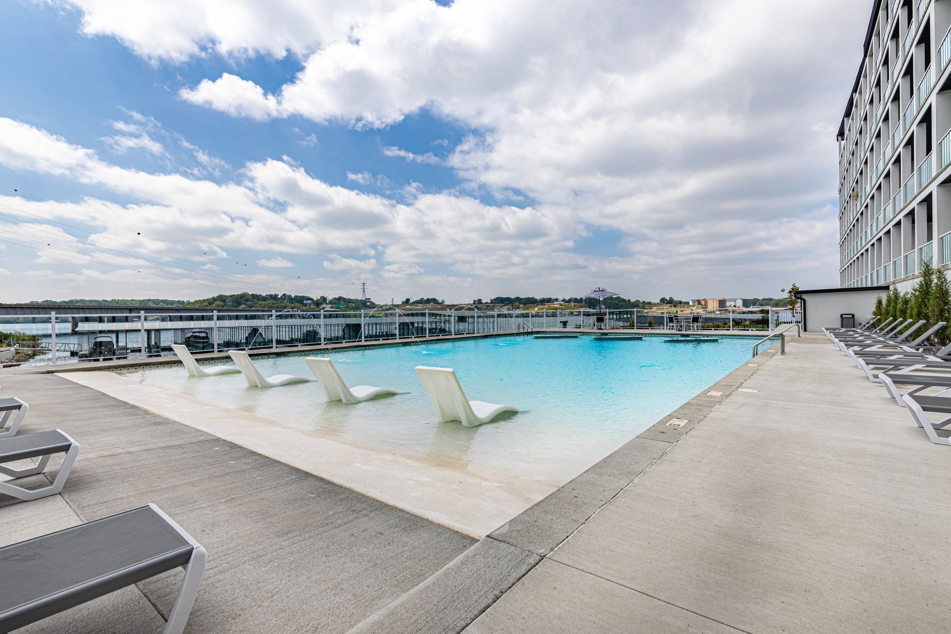 Rooftop pool with lounge chairs, blue water, and cloudy sky beside a modern building