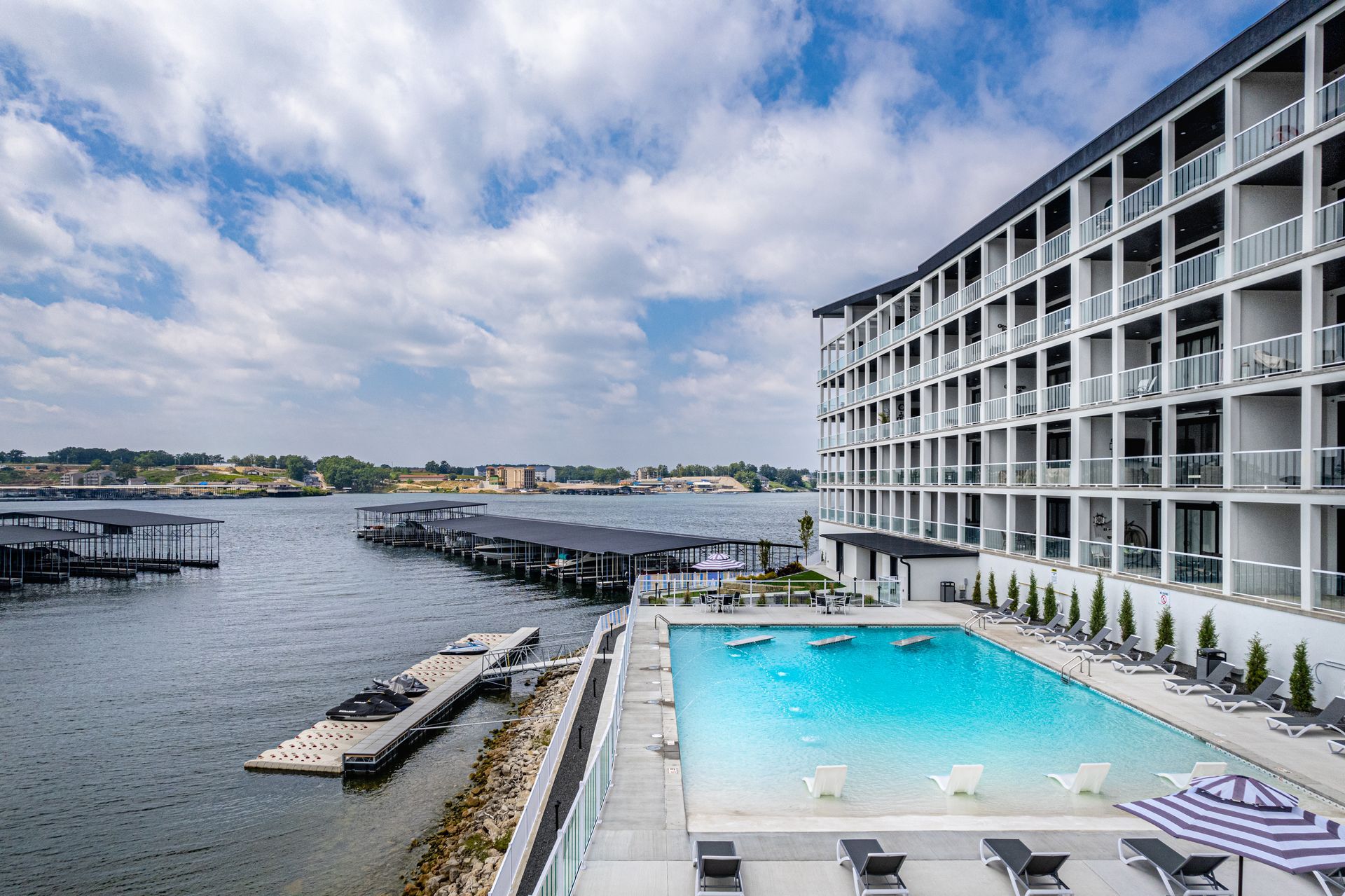 Waterfront hotel pool beside a modern building, overlooking a calm bay under a cloudy sky