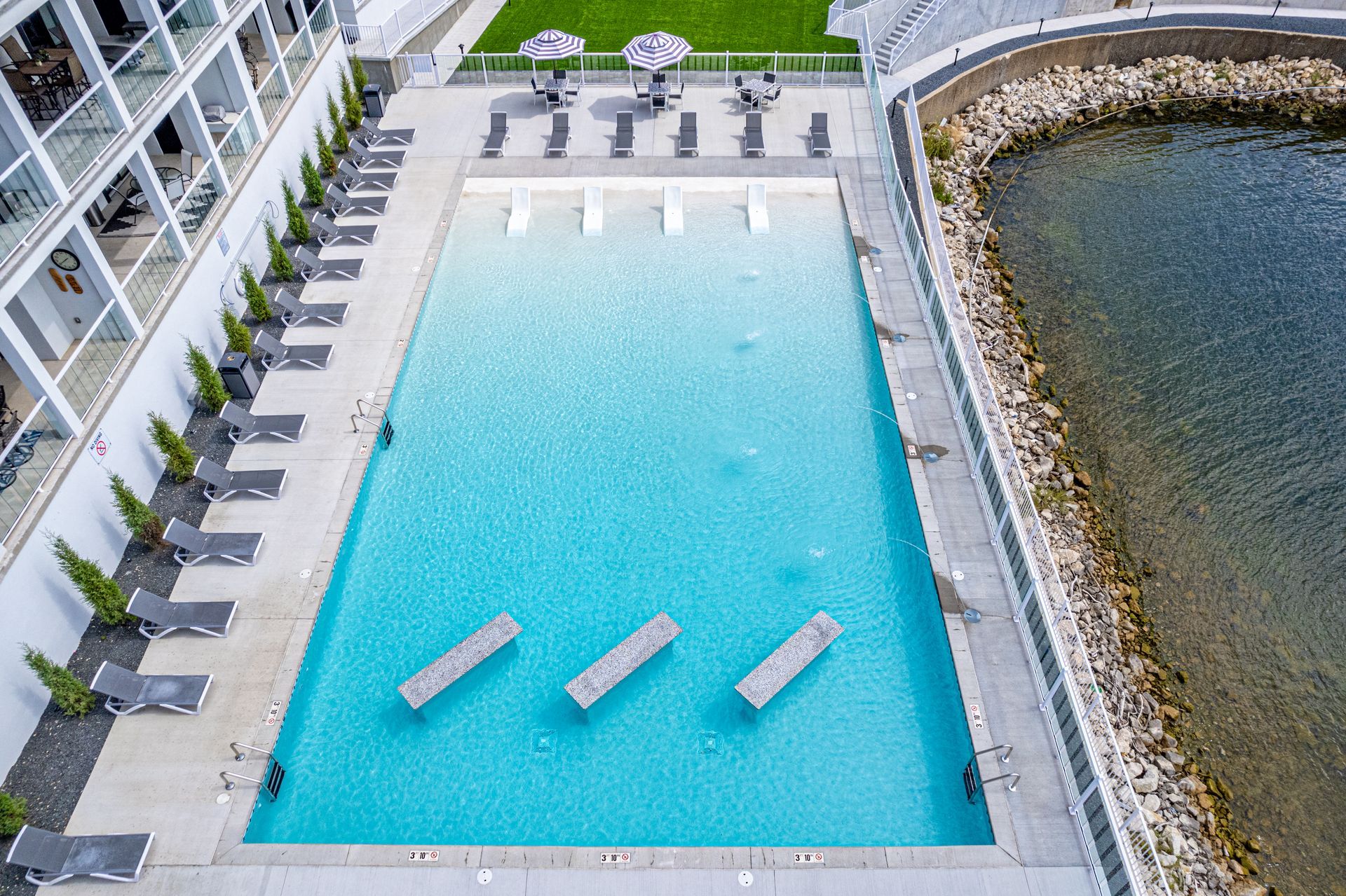 Rooftop pool with turquoise water beside a waterfront, lined with lounge chairs and greenery.