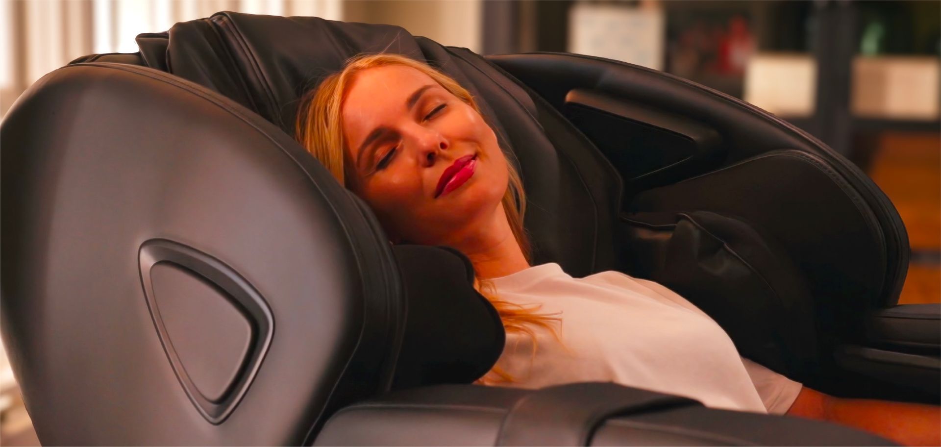 Person relaxing in a black massage chair, eyes closed, with warm orange lighting in the background