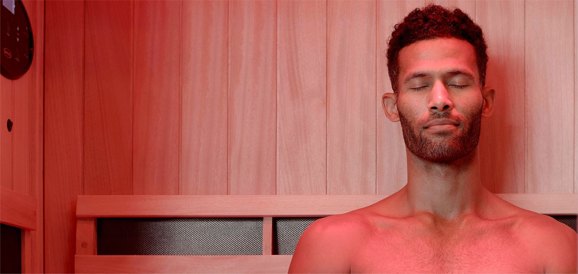 Man standing shirtless with eyes closed in a red-lit wooden sauna, leaning against the wall