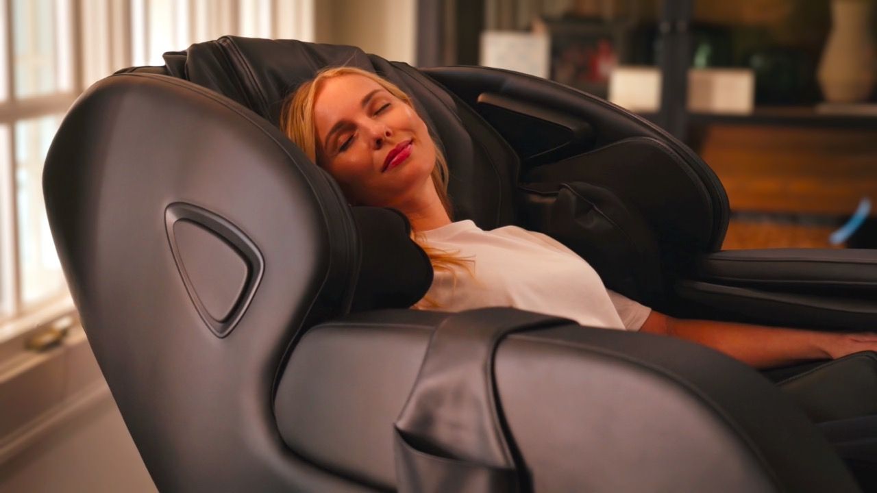 Person resting in a black massage chair, eyes closed, in a warmly lit room.