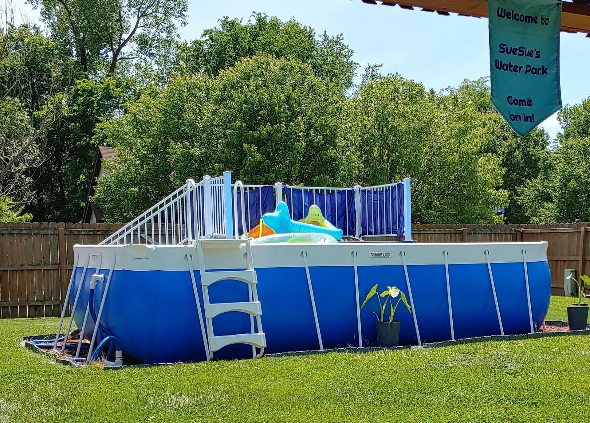 Blue above-ground pool in a grassy backyard with white ladder and slide, trees and fence in the background