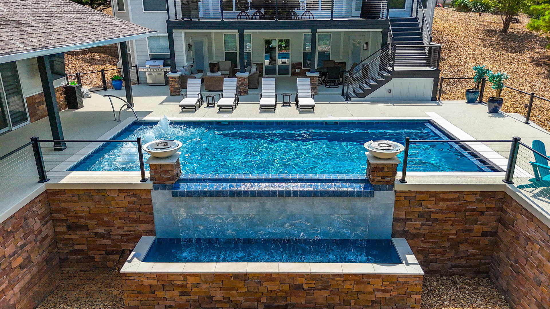 Modern pool with a waterfall, hot tub, lounge chairs, and a waterfront house in the background