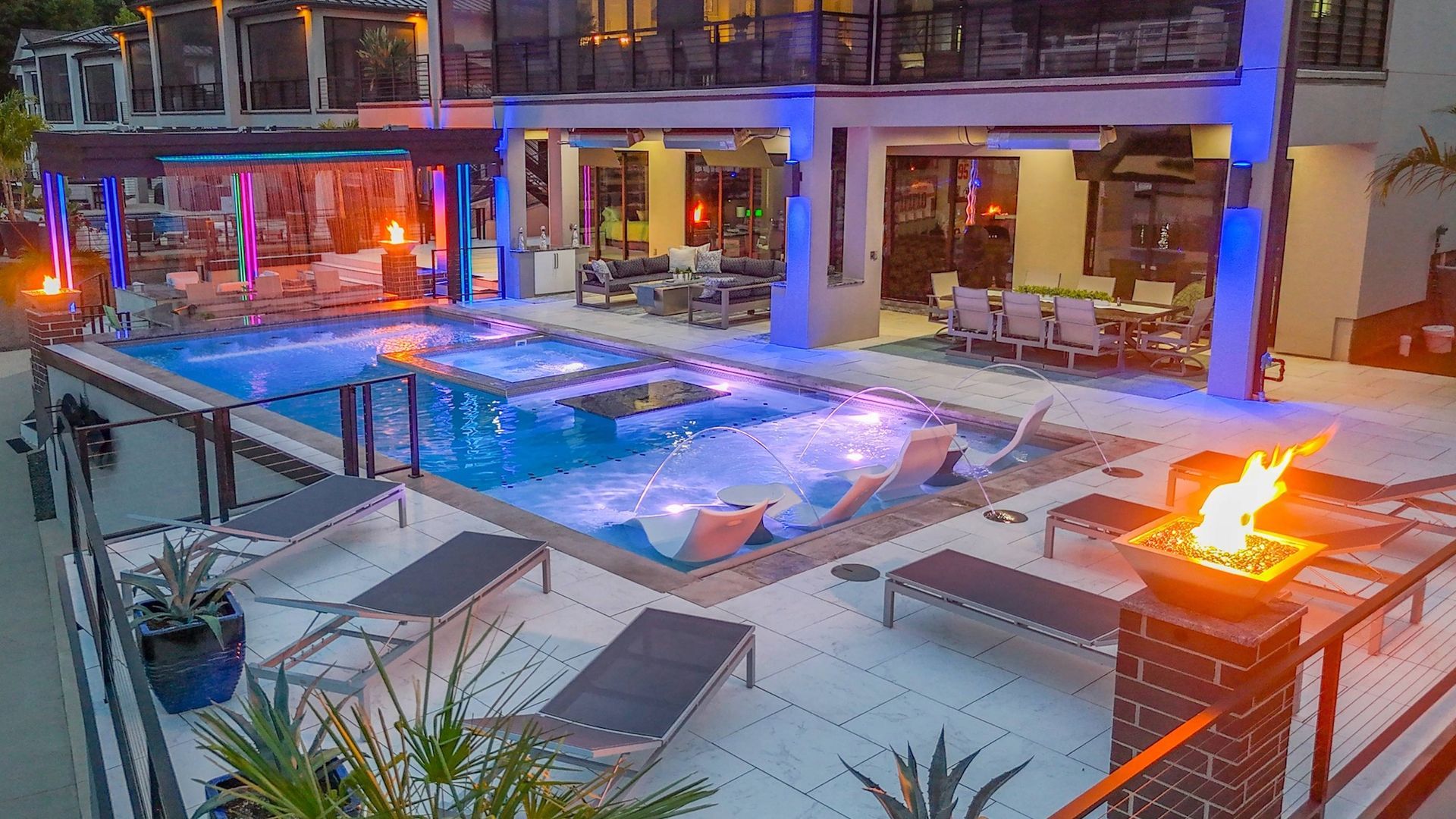 Nighttime pool courtyard with lit lounge chairs and a fire pit beside a modern building