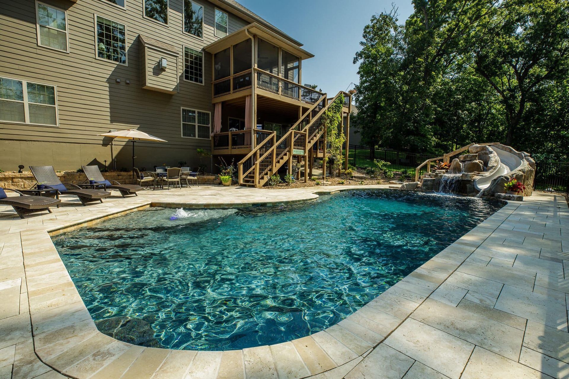 Backyard swimming pool beside a two-story house, with stone patio, stairs, and trees in the background