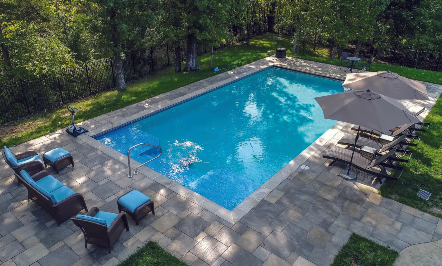 Backyard swimming pool with blue water, patio chairs, and a beige umbrella on a sunny day