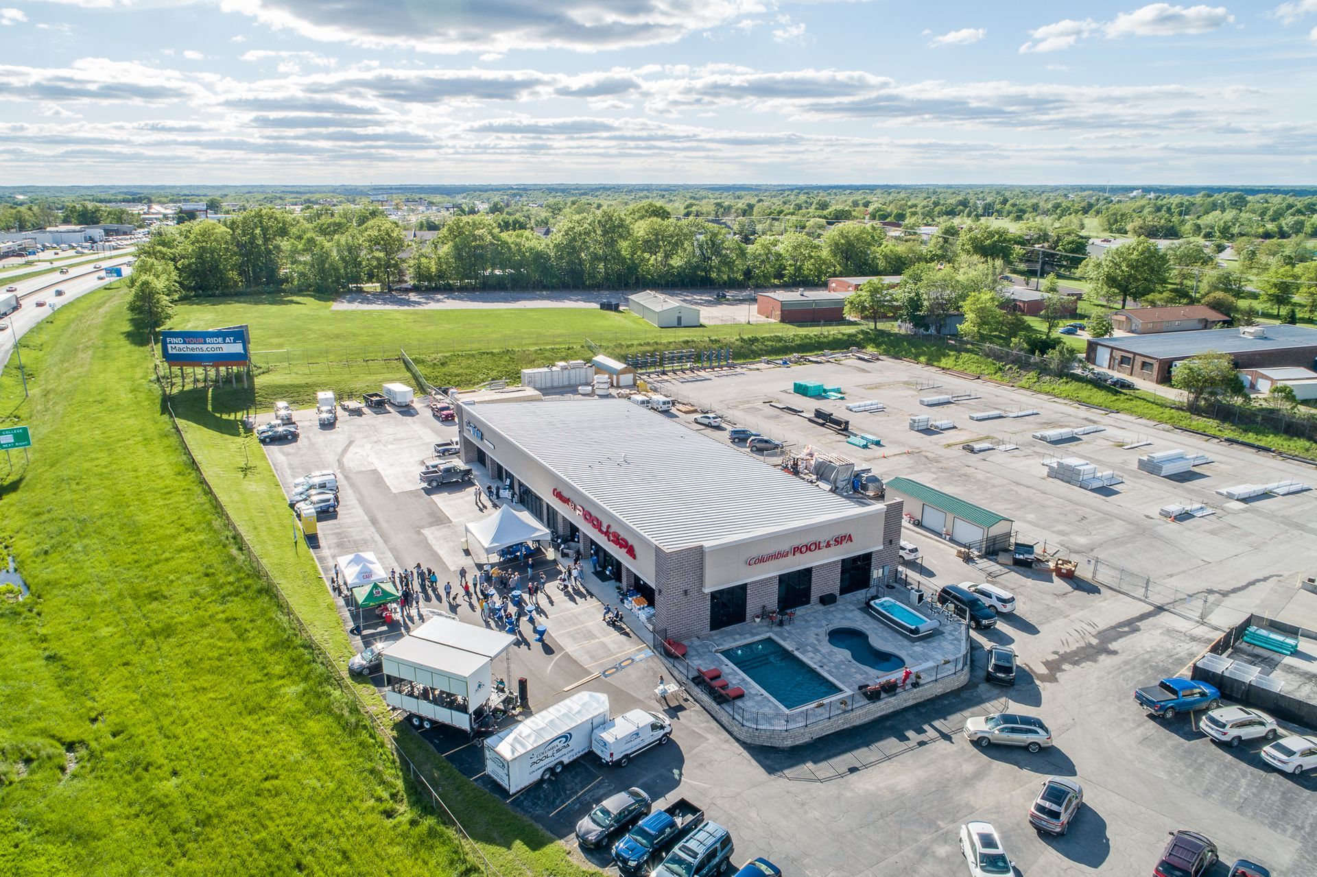 Aerial view of a car dealership with parked cars, a large striped roof, and surrounding green fields.