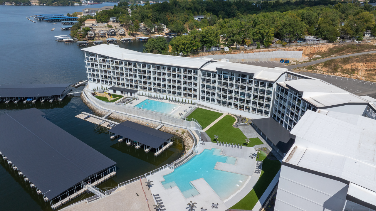 Aerial view of a modern waterfront hotel with a pool, dock, and boats along the shoreline.