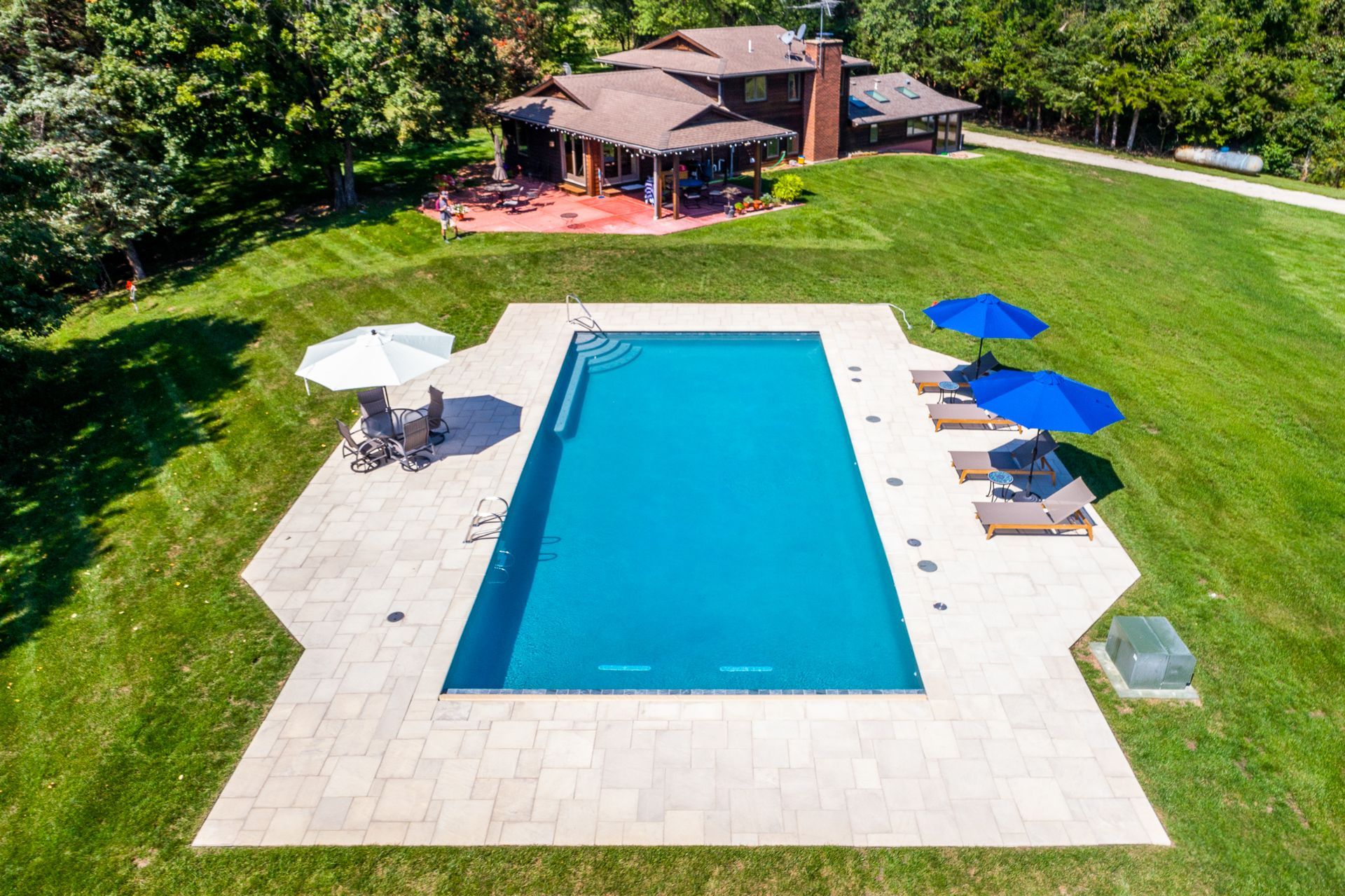 Aerial view of a backyard pool with blue water, lounge chairs, umbrellas, and a house surrounded by green grass
