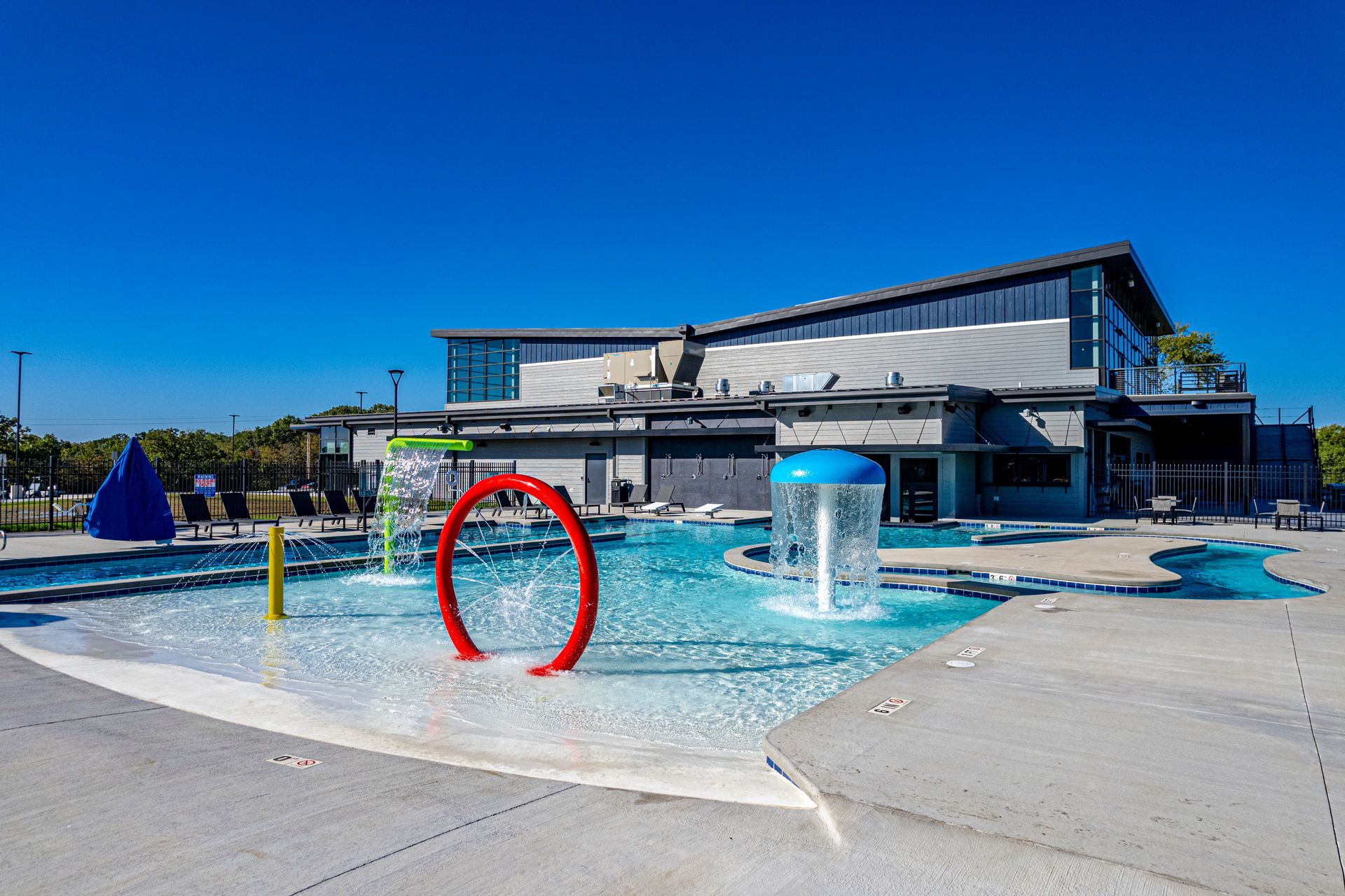 Outdoor splash pad with blue water jets, red ring, and modern building under a clear sky