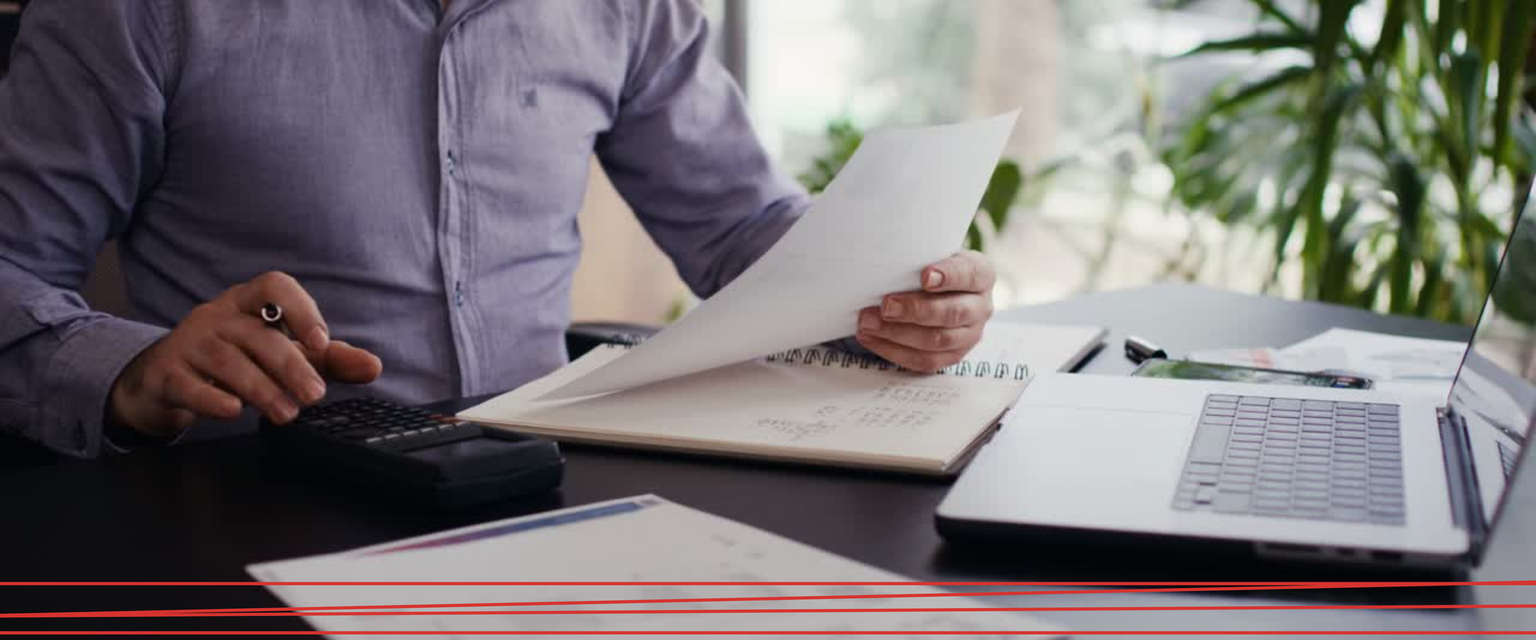 Una persona con una camicia bianca tiene in mano un documento vicino a un computer portatile e a una tazza di caffè su una scrivania scura.