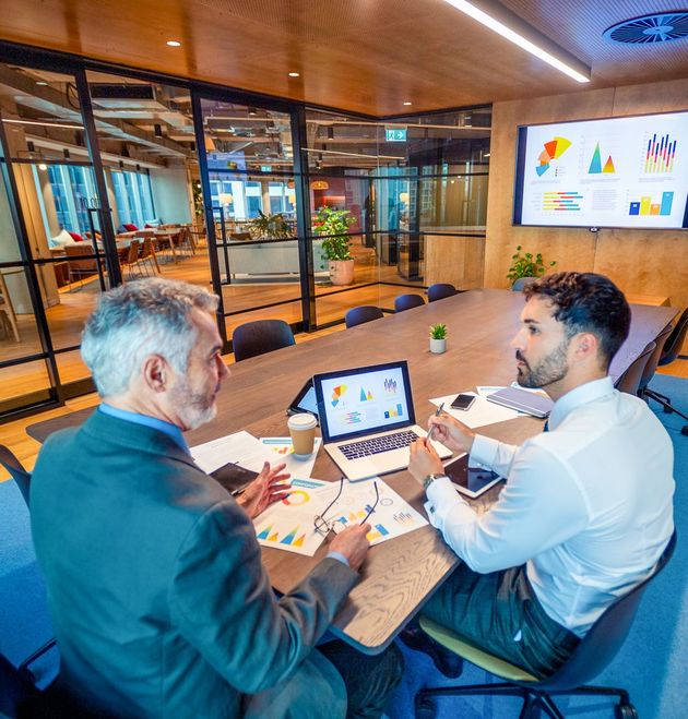 Business leader reviews financial information with a younger team member during a meeting in an office with glass walls.