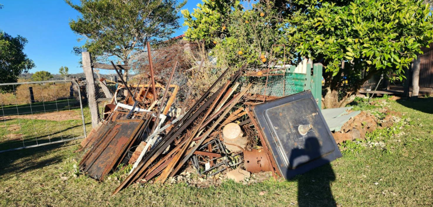 Pile of rusty metal and wood in a grassy yard under a blue sky, with a shadow in the foreground.