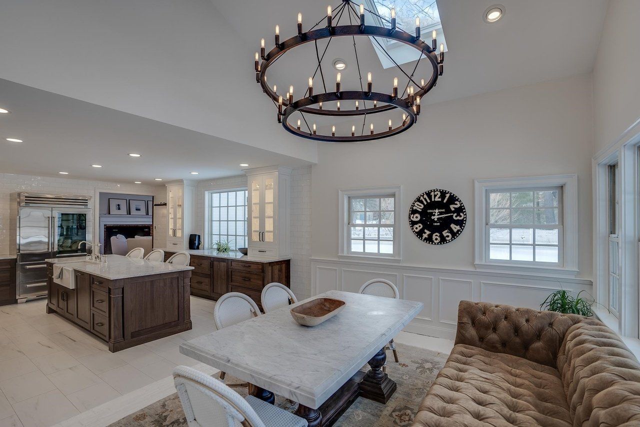 Bright open dining room and kitchen with white walls, dark chandelier, marble table, tufted bench, and island.