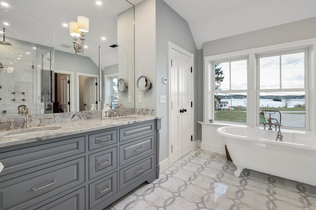 Bright bathroom with gray vanity, marble counters, freestanding tub, and large window overlooking water