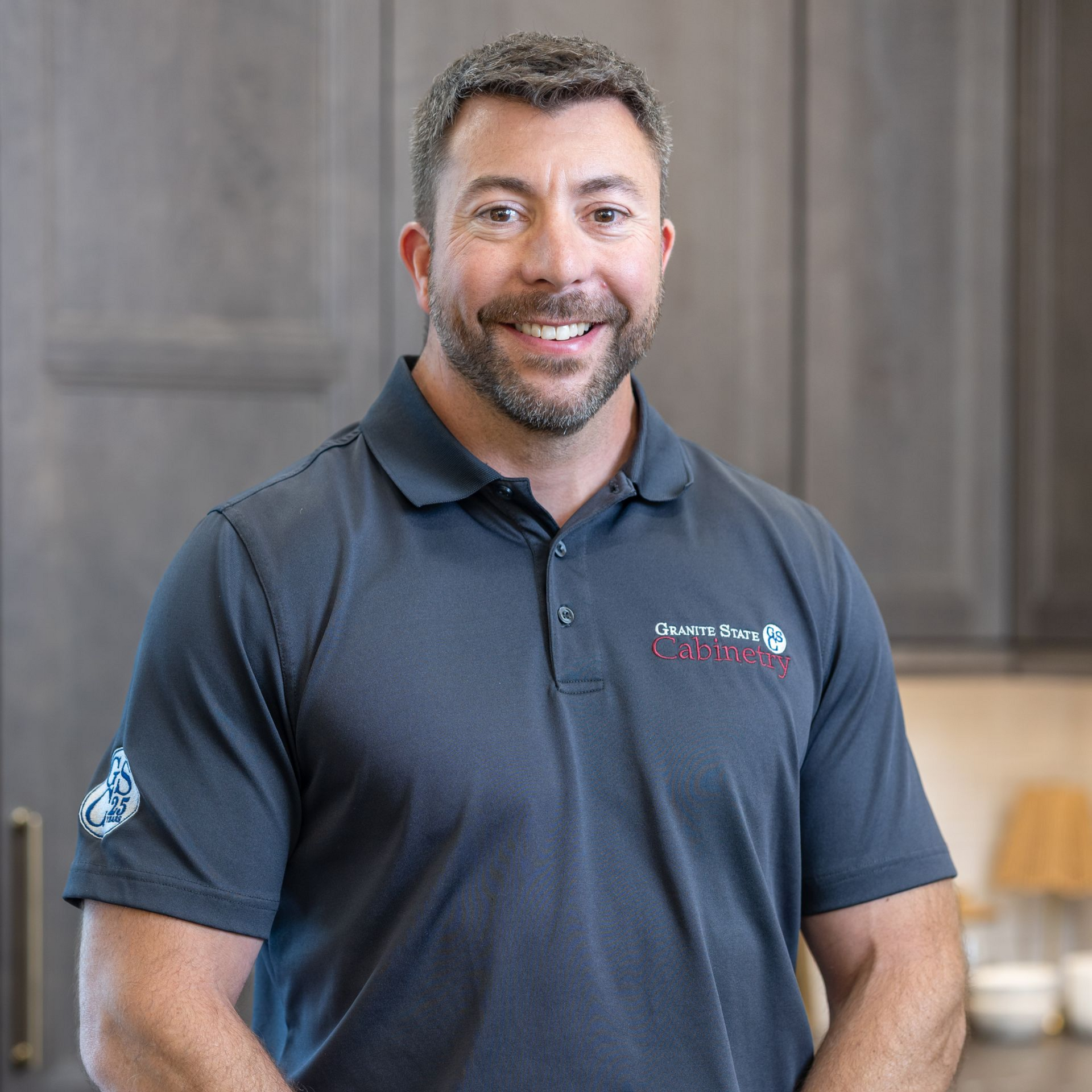 Smiling man in a dark polo shirt indoors, with white shelves in the background