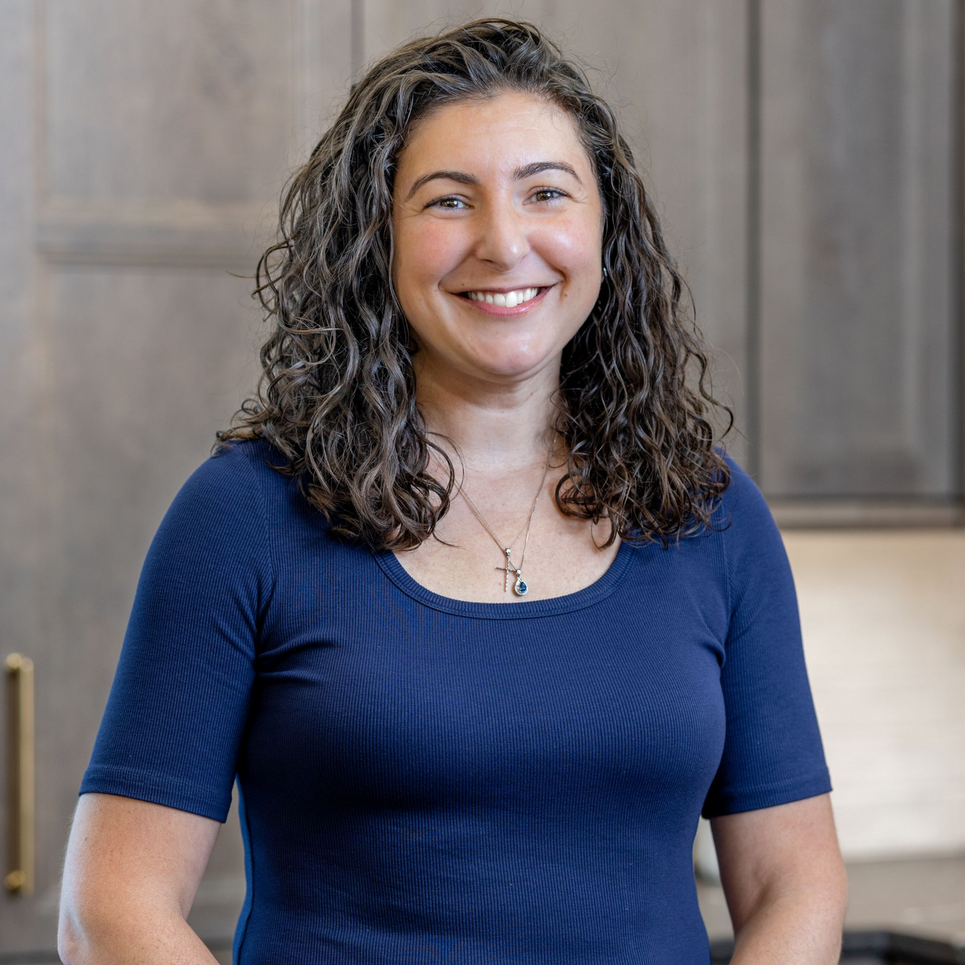 Smiling person with curly dark hair in a gray top, standing indoors near shelves.
