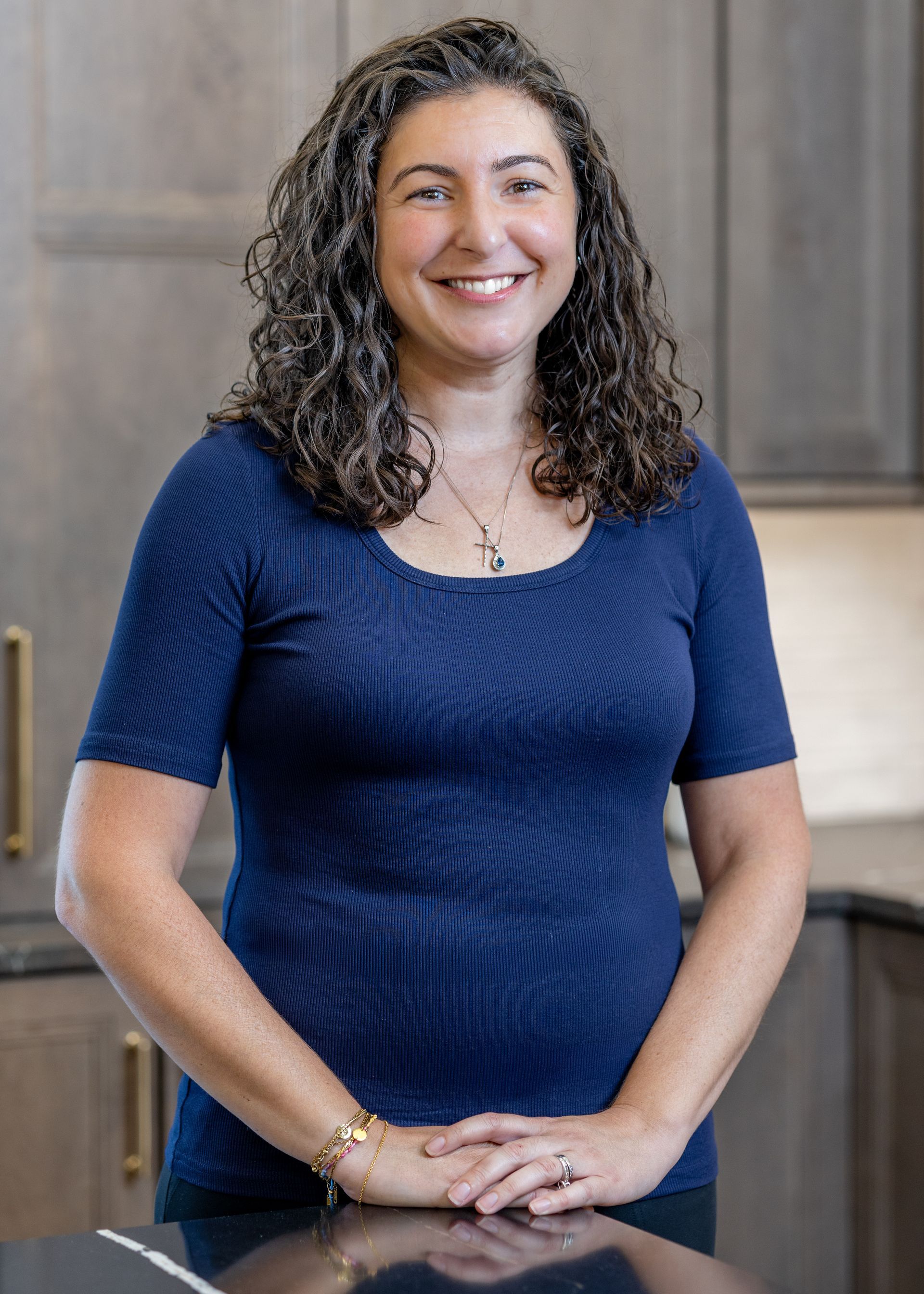 Smiling woman with curly hair standing in a bright kitchen with white cabinets and shelves.