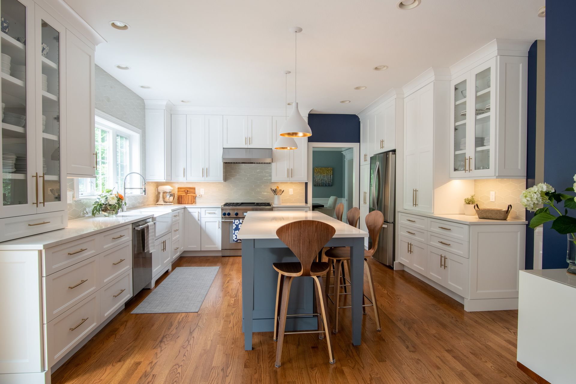 Bright white kitchen with blue island, wood floors, pendant lights, and a dining stool.