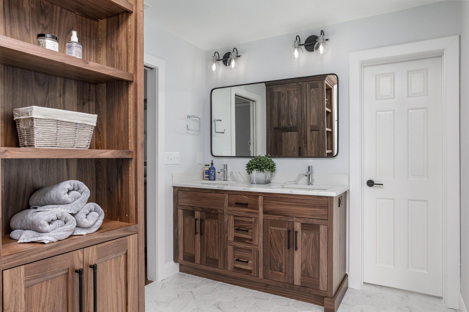 Modern bathroom with wood vanity, mirror, white tile floor, and shelving with rolled towels