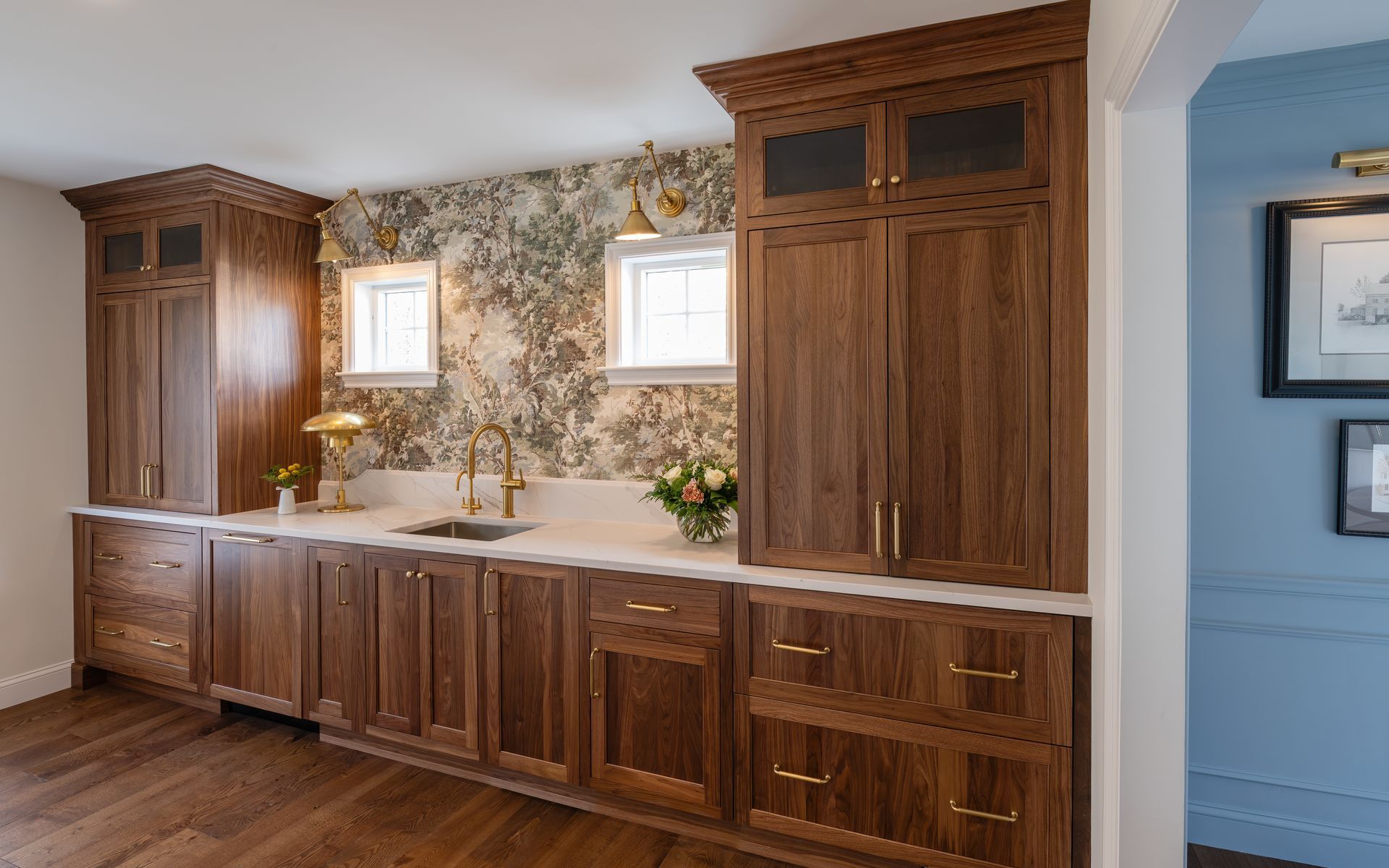 Wood-paneled kitchen wall with long cabinets, white countertops, floral backsplash, and two windows
